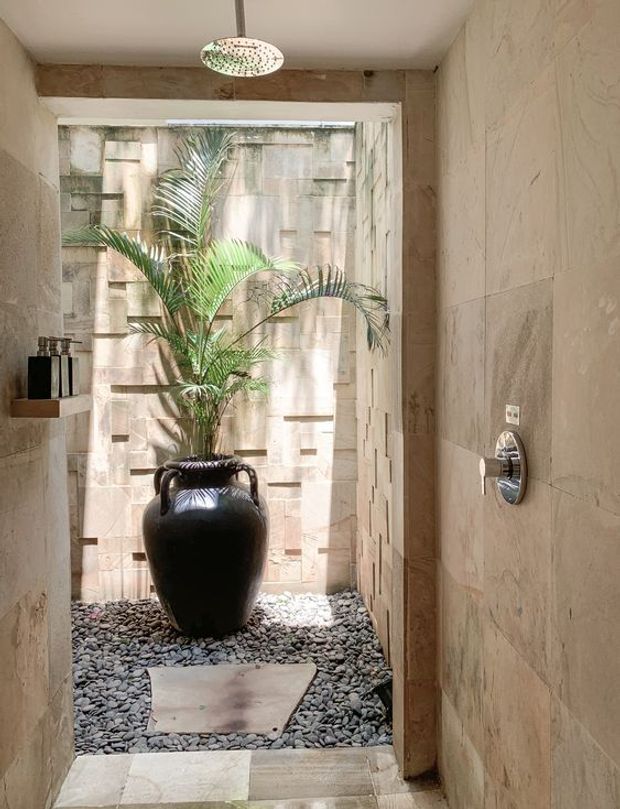 A compact, refined bathroom or wet-area passage opens onto a small internal courtyard. Large beige stone slabs line the walls and floor, with a chrome shower control on the right. The courtyard contains dark pebble landscaping, a stepping stone, a large black urn with a tropical palm, and a textured stone feature wall. A patterned pendant light hangs overhead, creating a warm, spa-like atmosphere.