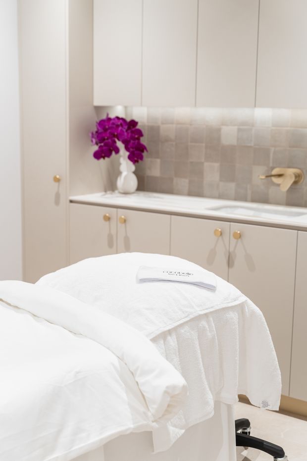Bright, refined treatment or wellness room with a white-covered massage table in the foreground. The room features pale built-in cabinetry with small brass-gold knobs, a white benchtop, a softly textured light neutral tiled splashback, and warm brass wall hardware. A white vase with vivid purple orchids adds the main accent colour.