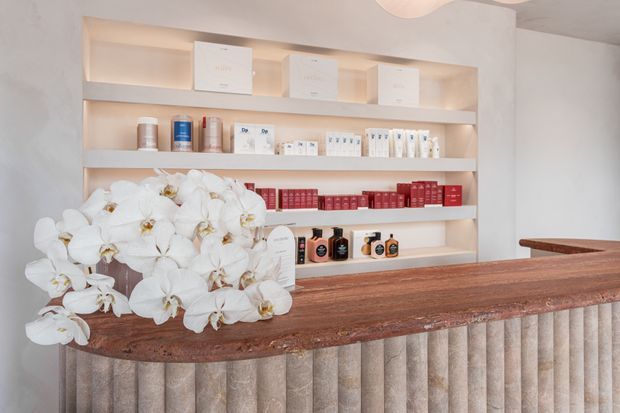 A refined reception counter in a softly lit retail or commercial interior. The foreground features a long reddish-brown stone or timber-look counter with a vertically fluted light-toned front, while illuminated open shelving behind displays skincare or beauty products and packaging. A large arrangement of white orchids provides a decorative focal point.