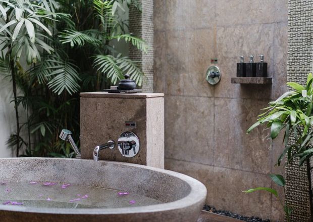A luxurious outdoor bathing area surrounded by dense tropical planting. The scene features a large rounded stone-look freestanding bathtub with rose petals, wall-mounted chrome shower controls and fixtures, a tiled shower wall, and a textured pebble-like accent wall. The earthy stone finishes and lush greenery create a resort-like atmosphere.