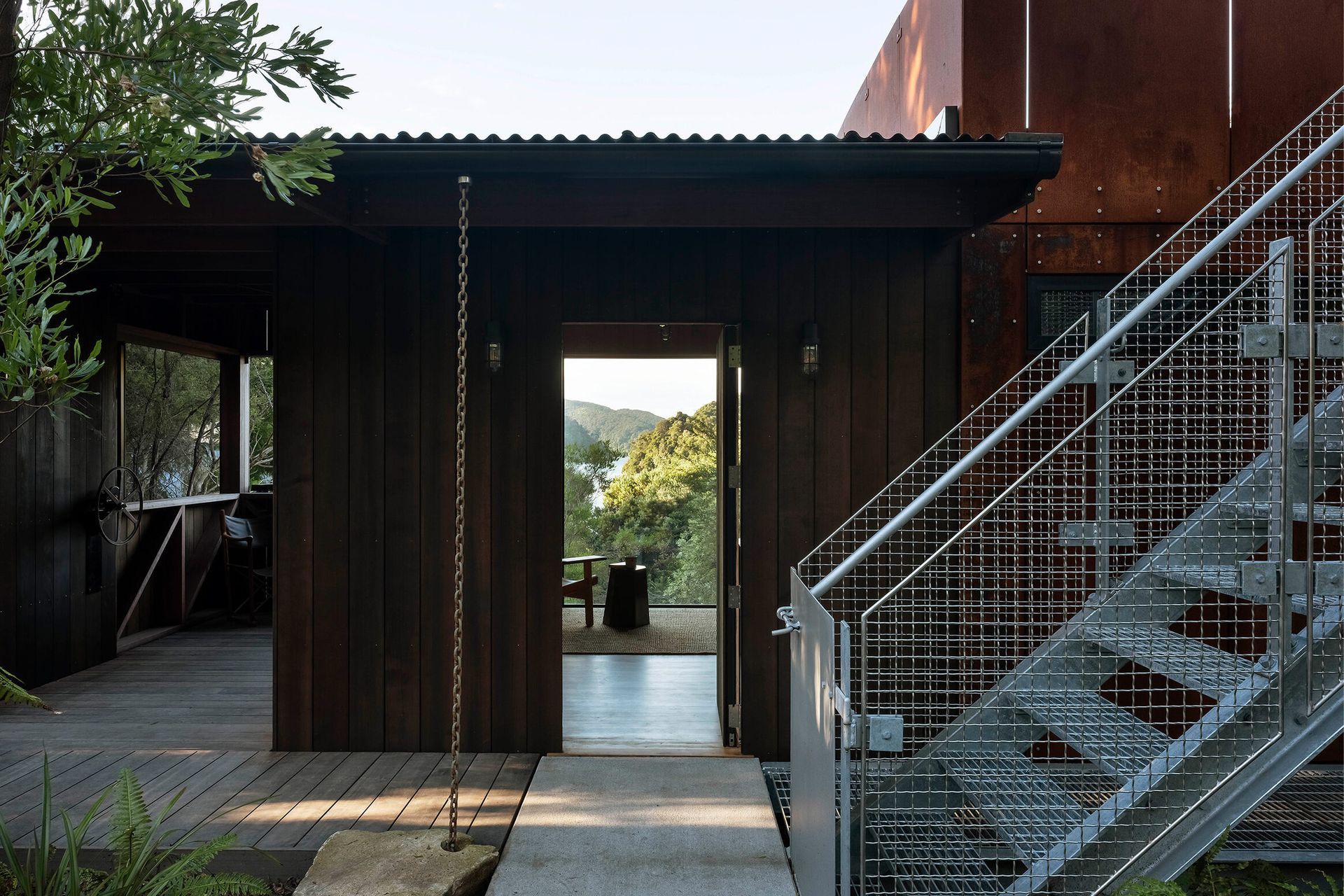 A metal staircase climbs to a treetop roof deck where the Cor-Ten cladding extends into a cantilevered balustrade.