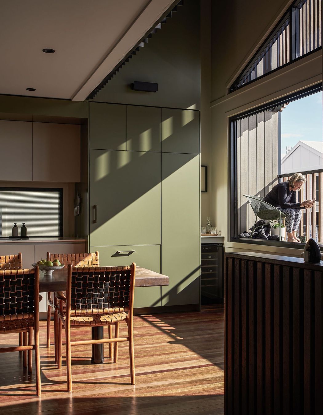 The palette features soft hues of sage and pale greens paired with natural timbers. To the right of the kitchen, a small balcony overlooks the street, capturing the morning and midday sun as it’s filtered through the timber batten screen.