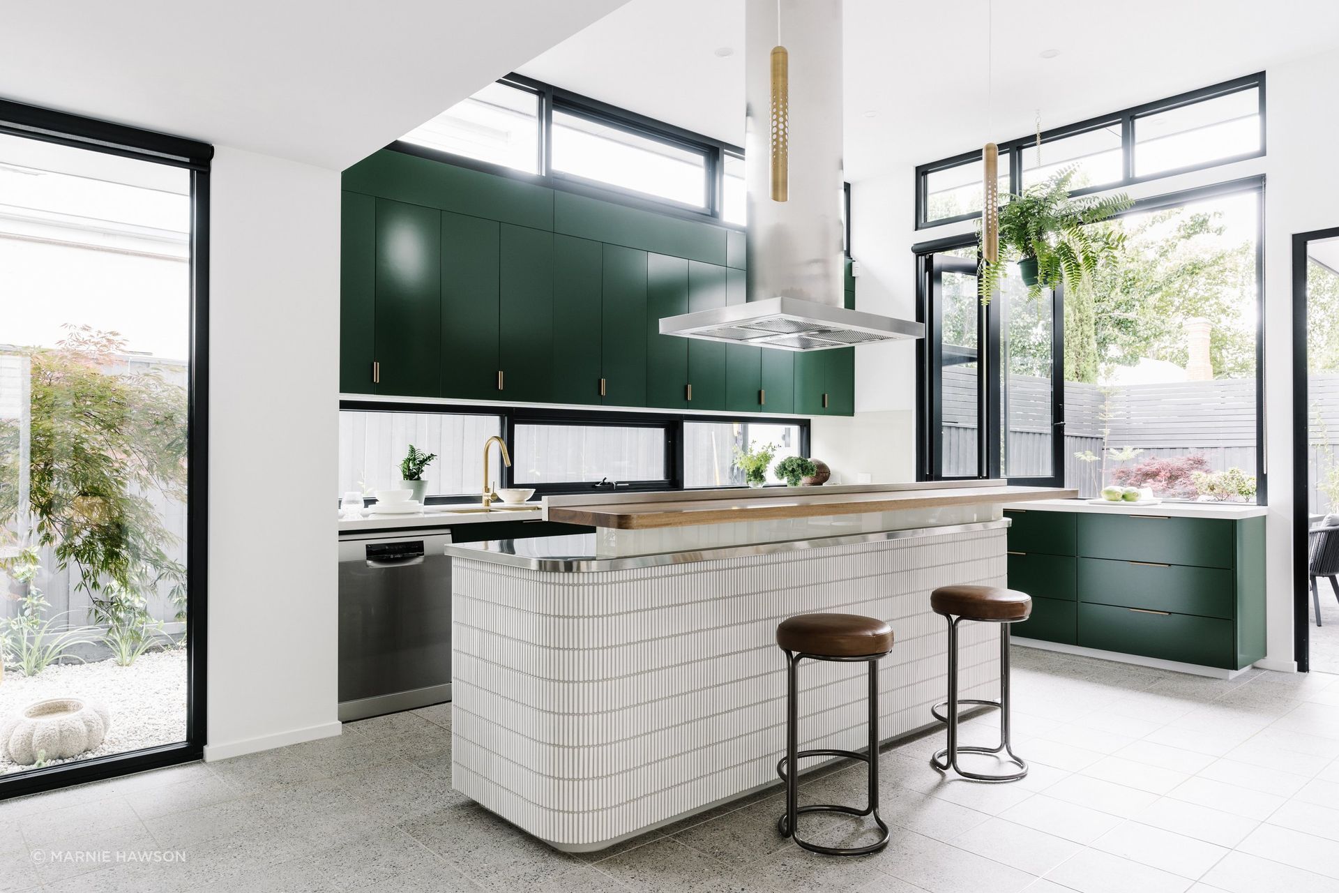 The island bench is clad in kit-kat tiles with brass light fittings above fitting with the rest of the home's mid-century modern details. A timber bar bench invites guests to sit, relax, and join in conversation while food preparation is underway.