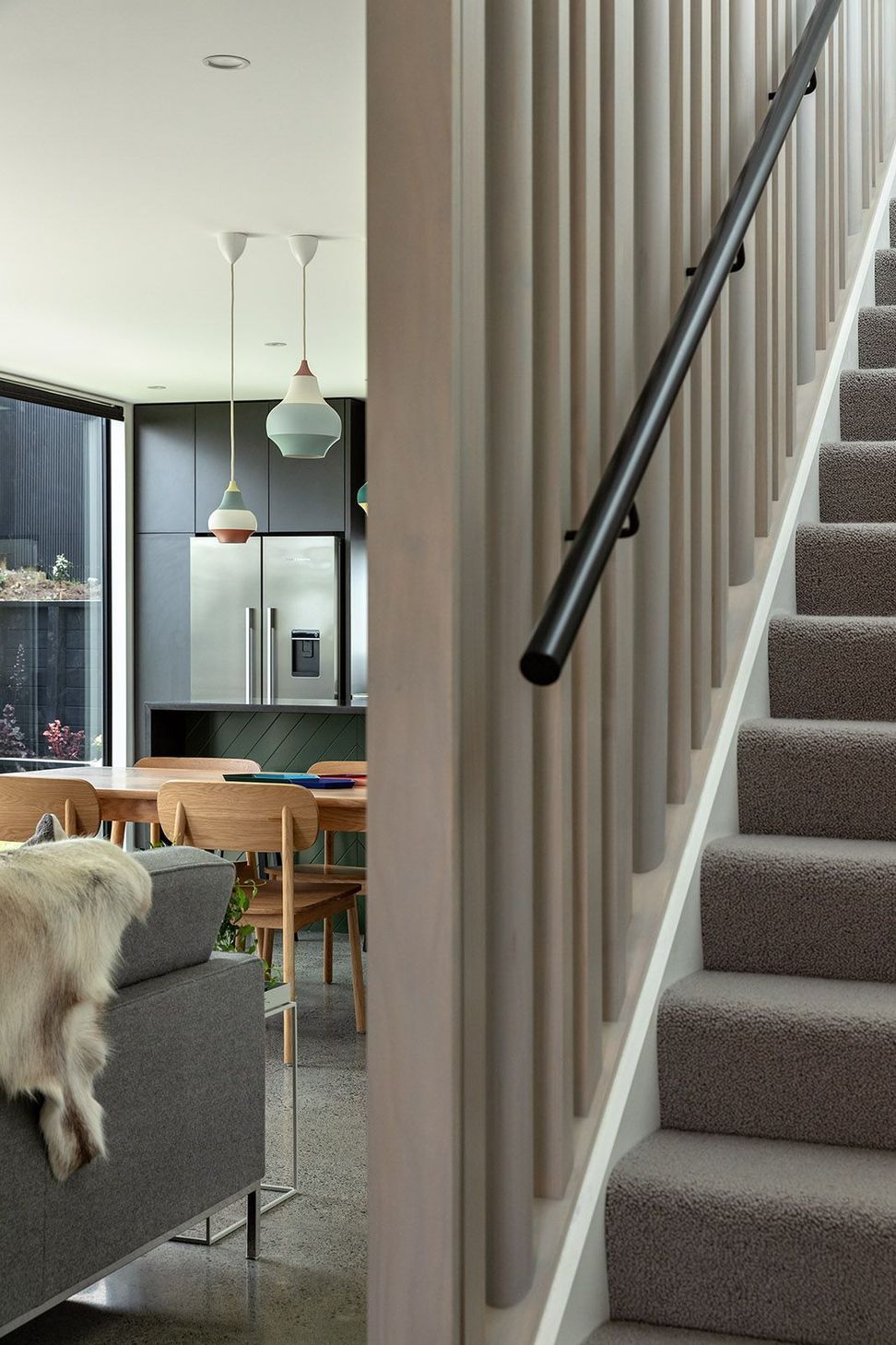 The custom staircase is screened by a grey-stained timber installation composed of varying timber profiles that reference Morse code. Beyond, the open-plan kitchen, dining and living spaces unfold beneath generous natural light.