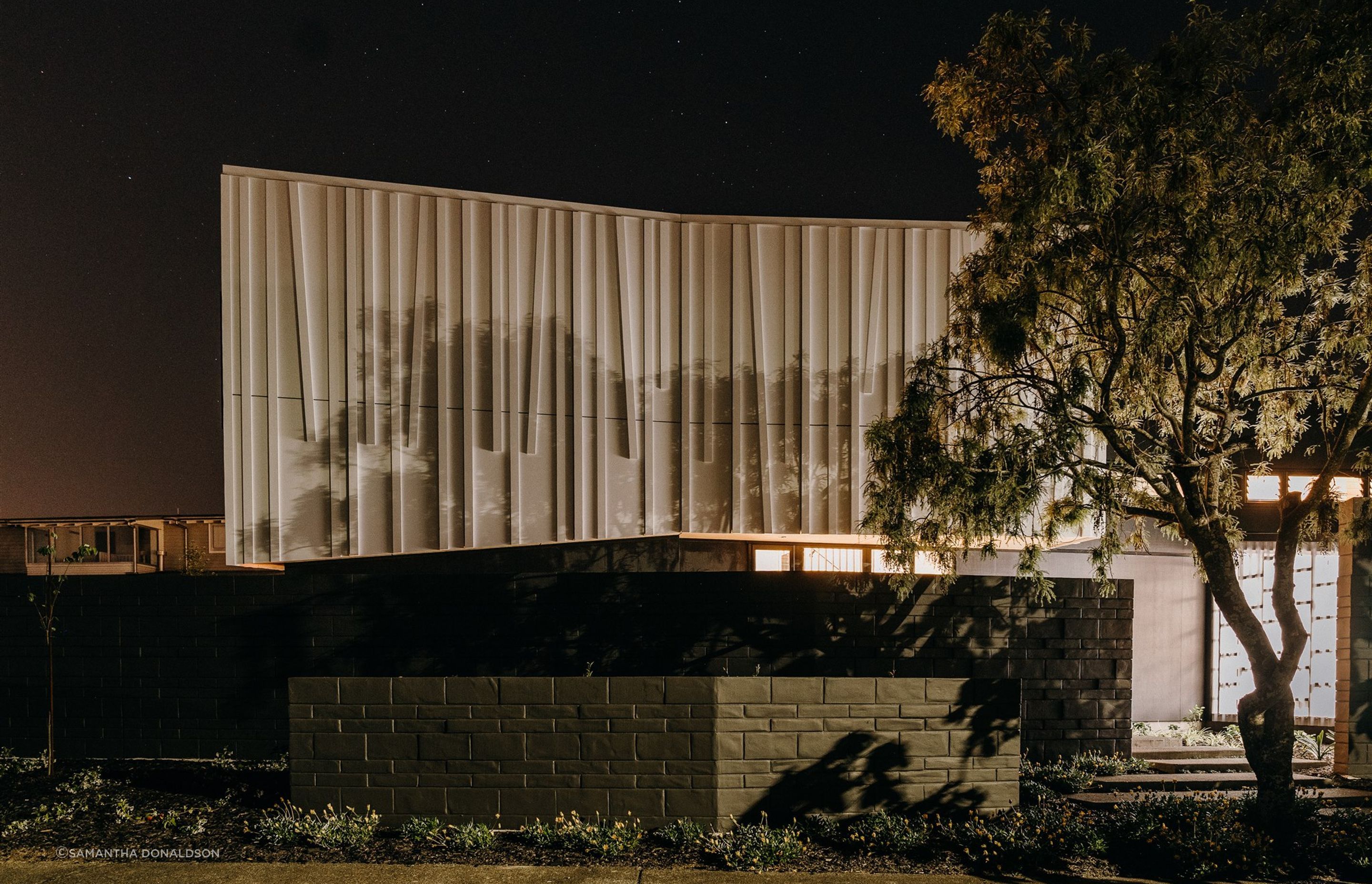 The 'shroud' or top level of the house is illuminated at night, with shadows cast from the surrounding trees to highlight the unique textural form. 
