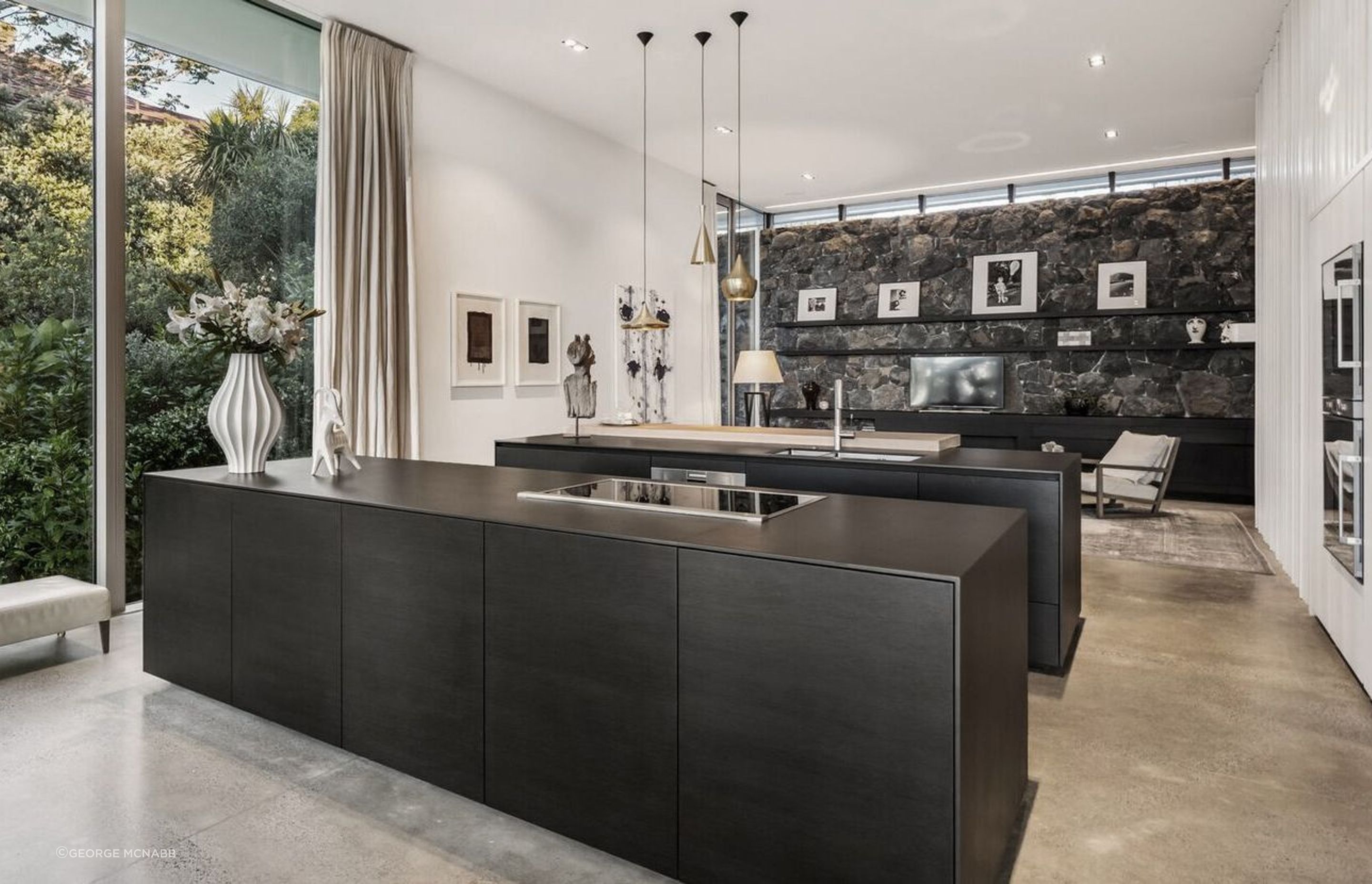 The view through the kitchen to the back of the house, where a basalt stone retaining wall sits underneath a clerestory window that draws morning light into the family space. 