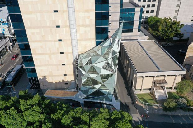 Aerial view of a dense urban commercial or institutional complex, featuring a prominent angular glass atrium or glazed entrance structure between taller buildings. The atrium has a faceted geometric steel-and-glass form, contrasting with pale stone or concrete facades, blue-tinted curtain-wall glazing, and a low ribbed metal-roof building. Tree-lined streets and surrounding urban buildings are visible.