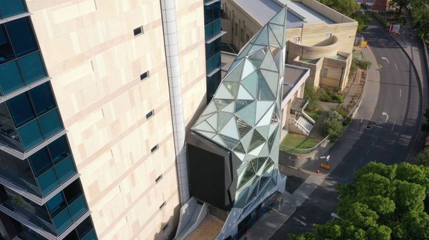 Elevated exterior view of a contemporary multi-storey urban building, featuring a prominent angular glass-and-metal faceted atrium or entrance structure between larger glazed and pale panel-clad volumes. Curved roof forms, terraces, access roads and landscaped edges are visible around the complex.