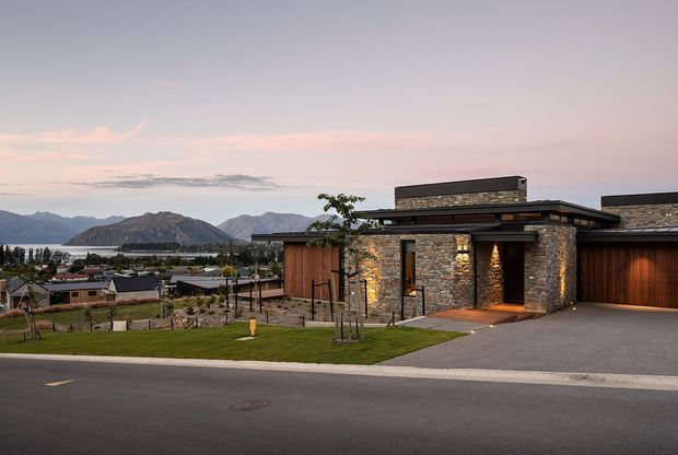 Contemporary single-storey residence viewed from the street at dusk, featuring a low flat roof, dark horizontal roof planes, warm timber-clad sections, and prominent rough stone veneer walls with integrated exterior lighting. A broad driveway and landscaped front lawn lead to the entry and garage. Mountains and a settled neighbourhood are visible in the background.