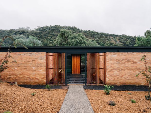 Single-storey low-profile building facade set against a wooded hillside. The exterior combines warm exposed brick walls with a dark flat roof edge, a central timber entry door, and vertical timber screening flanking the recessed entrance. A straight concrete or aggregate path leads through a sparsely planted, mulched landscape.
