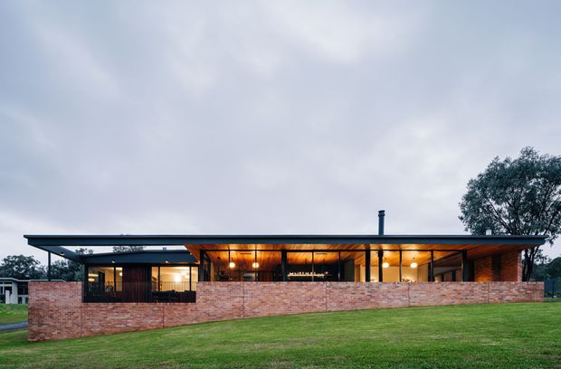 Low, single-storey contemporary house viewed from the exterior across a lawn. The building has a broad flat roof with deep overhangs, extensive glazing, dark slender framing, warm timber-lined interior ceilings visible through the openings, and a long exposed brick boundary wall. A chimney projects above the roof, with mature trees and neighbouring buildings in the background.