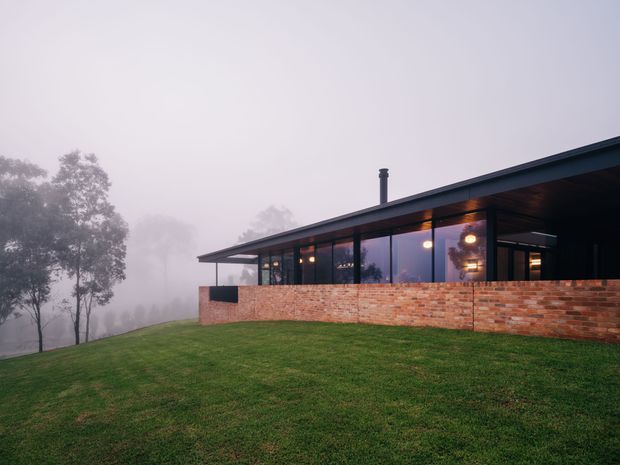 A low, modern single-storey house set on a sloping grassy site in misty, wooded surroundings. The building has a long flat roof with broad overhangs, extensive dark-framed glazing, and a prominent red brick base and wall. Warm interior lighting glows through the windows, creating a strong contrast with the cool foggy exterior.