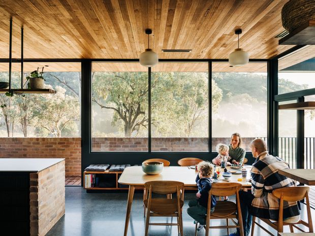 A warm, family-oriented dining and kitchen area with a timber-lined ceiling, exposed brickwork, dark-framed floor-to-ceiling glazing, and a timber dining table surrounded by chairs. A kitchen island is visible at left, while large windows overlook a leafy landscape. Pendant lights and built-in bench seating complete the relaxed contemporary interior.
