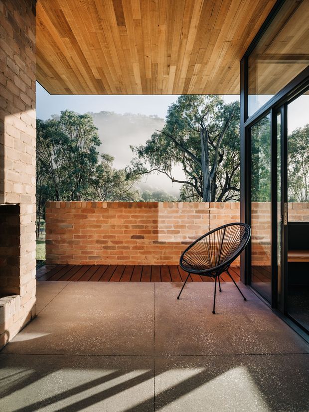 A sheltered outdoor terrace or balcony framed by exposed brick walls and large black-framed glazed sliding doors. The terrace combines a warm timber-lined soffit and timber decking strip with a textured concrete floor, a low red-brick parapet, and a single black woven lounge chair. Eucalyptus trees and a hazy landscape are visible beyond the parapet, creating a warm, nature-connected architectural setting.