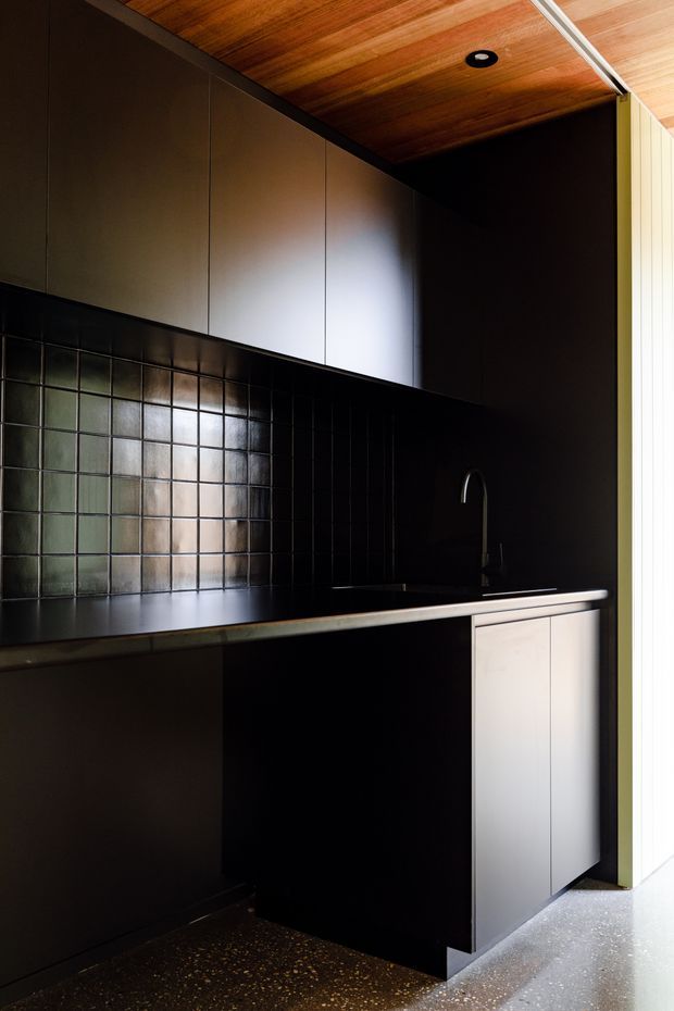 Compact, dark-toned kitchen or kitchenette with matte black cabinetry, a black benchtop and glossy square tiled splashback. A slim undermount sink with curved tap sits beneath upper flat-panel cupboards. The ceiling is lined with warm timber boards and includes a recessed downlight, while bright vertical glazing or a light-coloured opening appears at the right edge. The space has a refined, moody contemporary character.