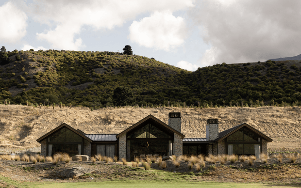 Low, single-storey lodge-style residence set into a dry, hilly landscape. The building has a symmetrical multi-wing composition with dark timber gable fronts, stone-clad walls and chimneys, dark metal roofing, and large glazed openings. Native grasses and rugged terrain surround the home, with wooded hills rising behind it under a partly cloudy sky.