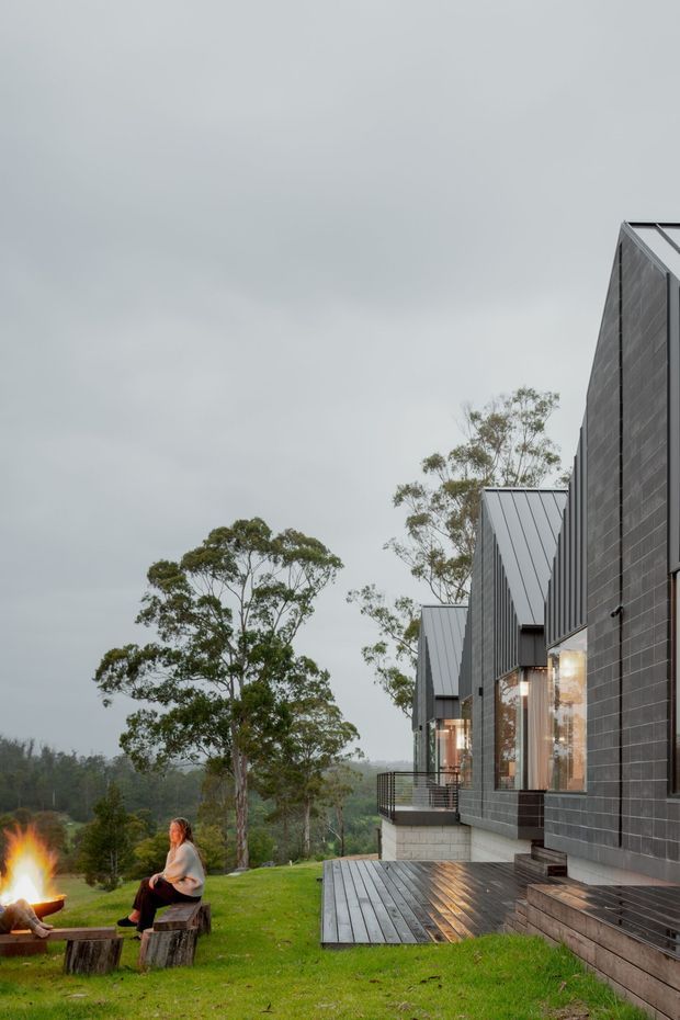 Exterior view of a contemporary dark-clad multi-level building set in a green, wooded landscape. The facade features dark rectangular cladding, tall glazed openings, steep metal gable roofs, balconies, and a broad timber deck. A fire pit and seating occupy the lawn in the foreground.