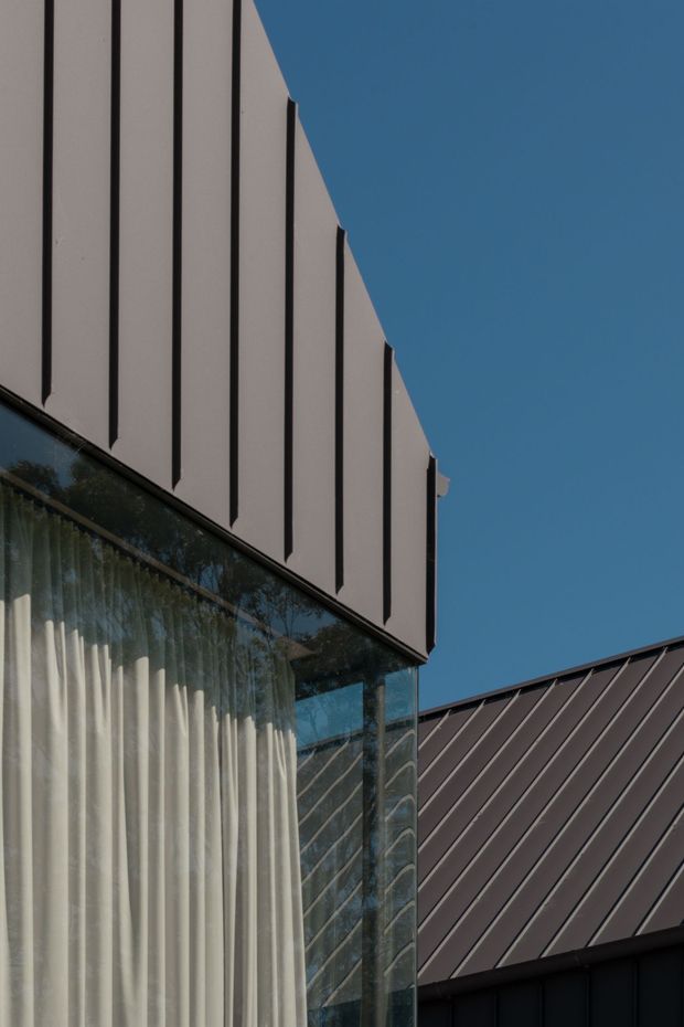 Close-up architectural photograph of a contemporary building facade against a clear blue sky. The image highlights pale vertical facade panels with dark recessed seams, a glazed corner with sheer curtains, and a contrasting dark standing-seam metal roof.