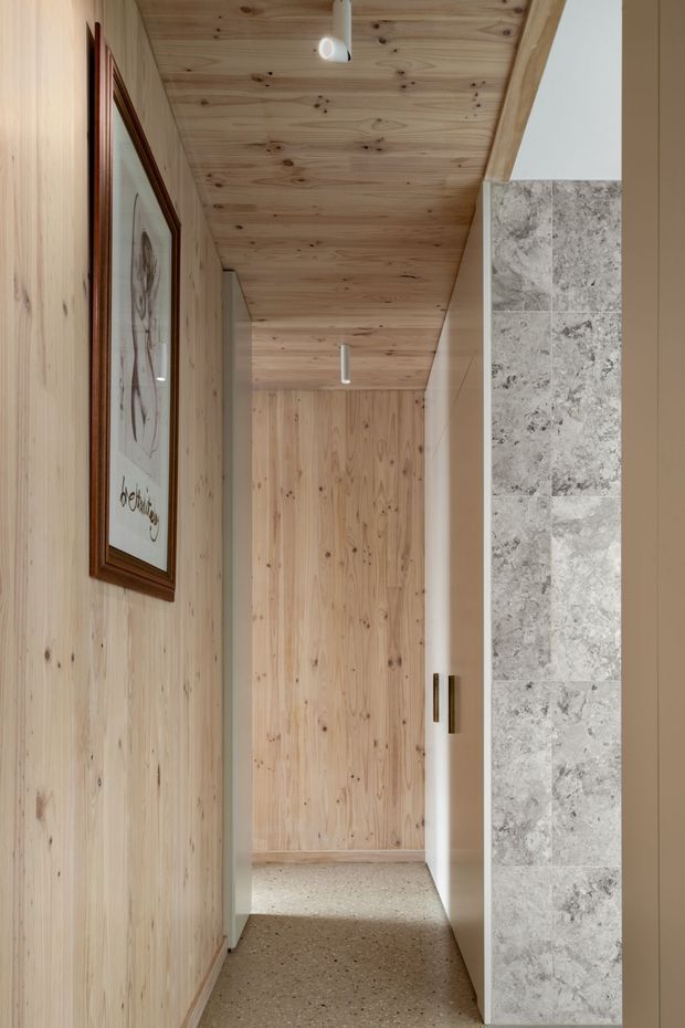 A narrow, refined hallway lined with pale timber wall and ceiling panelling, leading to a timber door or built-in joinery at the far end. A terrazzo-like floor and a vertical strip of grey stone tile frame the right side, with slim recessed or surface-mounted cylindrical lights and framed artwork adding subtle detail.