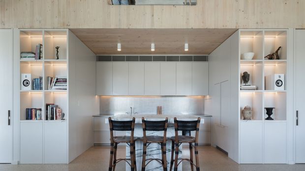 A compact contemporary kitchen and dining area integrated into built-in cabinetry. A central island with seating for four sits beneath a timber-lined recessed ceiling, with pale cabinetry, stainless-steel splashback, open display shelving, books, and concealed storage on both sides.