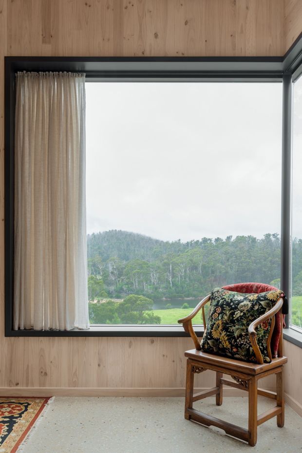 A quiet interior corner with warm vertical timber wall panelling, a large black-framed corner window overlooking a lush wooded landscape, sheer beige curtains, and a carved wooden armchair upholstered with a dark floral cushion. The pale terrazzo-like floor and natural materials create a calm, refined atmosphere.