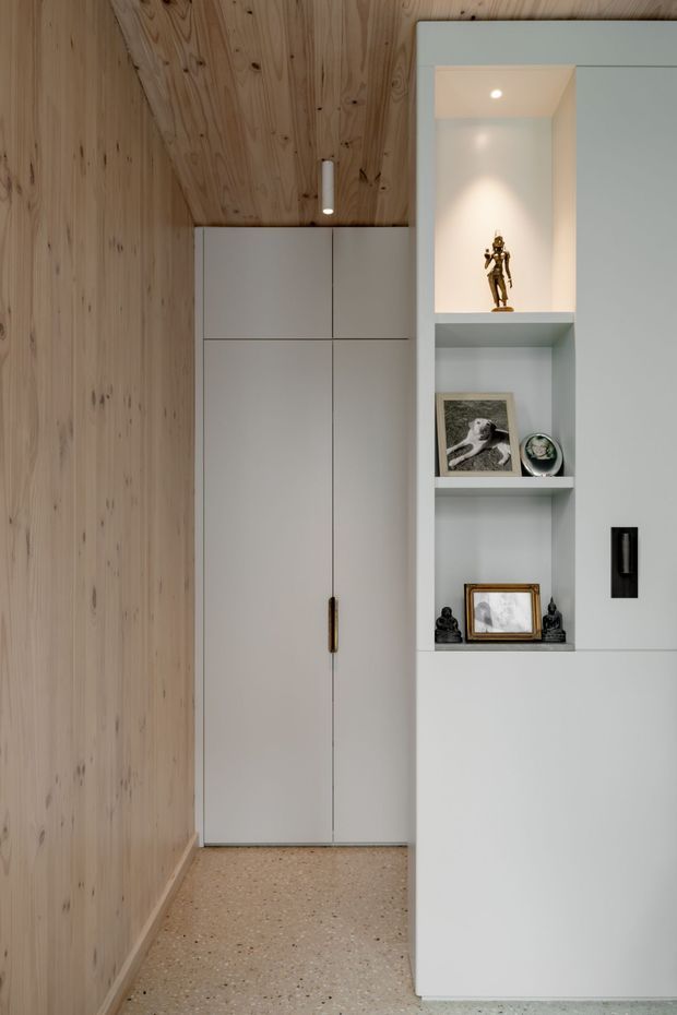 A compact, carefully detailed interior corner featuring pale natural timber wall and ceiling panelling, a flush white built-in wardrobe, and a tall white display cabinet with recessed illuminated niches. Small artworks and sculptural objects are arranged on the open shelves. The floor is a light terrazzo-like finish, and the overall composition is clean, restrained, and warm.