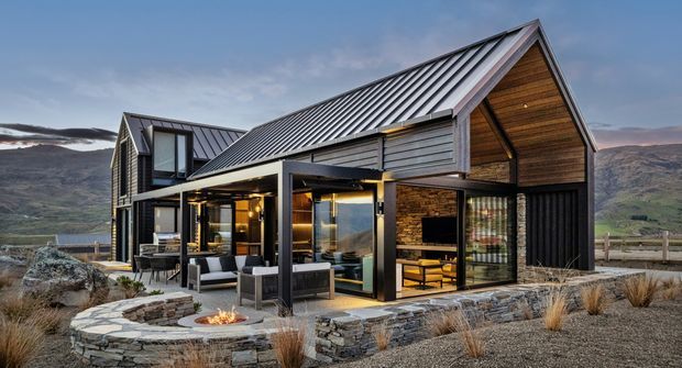 Exterior view of a contemporary mountain home with steep gabled metal roofing, dark vertical cladding, timber-lined gable soffits, extensive glazing, and stone detailing. The house opens onto a paved terrace with outdoor seating and a built-in fire pit, set against a rugged alpine landscape.