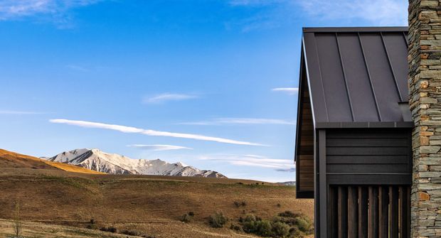 Exterior architectural detail of a dark metal-clad structure with a steeply sloped roof and vertical timber-like screening, joined to a rough stacked-stone wall. The building sits in an open alpine or high-country landscape, with rugged snow-dusted mountains visible in the background beneath a clear blue sky.