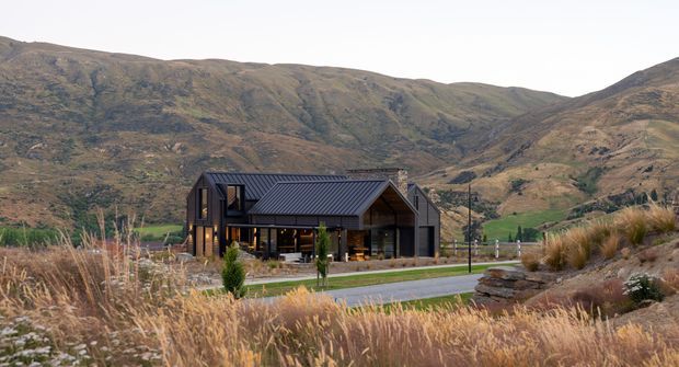 A contemporary mountain home set within a rugged alpine valley landscape. The dark metal pitched roof, timber-and-glass facade, and warm interior lighting contrast with the surrounding dry grasses, landscaped foreground, and steep brown hillsides.