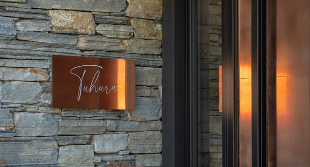 Close-up of a sophisticated building entrance featuring a dark grey stacked-stone wall, a dark-framed glazed doorway, and a rust-toned metal entry panel. A rectangular rust-coloured sign with elegant script lettering is mounted on the stone wall, while warm lighting reflects in the glazing and highlights the metal surface.