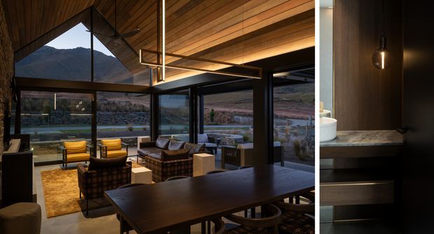 A refined open-plan living and dining space with extensive floor-to-ceiling glazing overlooking a rugged mountain and valley landscape. The room features a large dark timber dining table, lounge seating, yellow accent armchairs, dark framed glazing, a timber-lined ceiling with integrated linear lighting, and a stone-textured feature wall. A narrow adjoining view shows dark timber built-in joinery with a stone benchtop and pendant light.