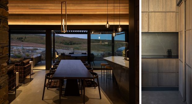 A warm, contemporary open-plan dining and kitchen interior with a long dining table, woven dining chairs, pendant lighting, dark cabinetry, and a stone kitchen island. Full-height glazing opens views toward a rugged landscaped outdoor setting. A timber-lined ceiling and exposed stone wall add natural texture, while the right side shows a compact integrated kitchen or utility joinery zone.