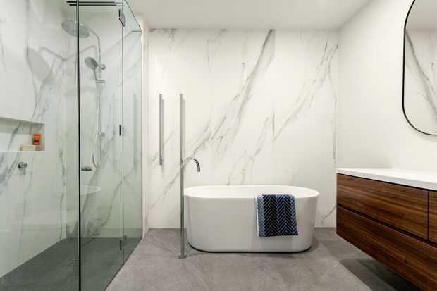 Contemporary luxury bathroom with a freestanding white bathtub, frameless glass walk-in shower, large-format white marble-look wall slabs with grey veining, grey floor tiles, and a floating dark timber vanity with a white benchtop. Brushed metal tapware and minimal black-edged mirror detailing add contrast.