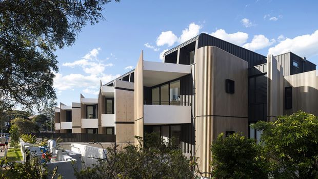 A contemporary multi-unit residential development viewed from the landscaped street frontage. The building comprises a series of stepped, articulated volumes with dark standing-seam or corrugated metal upper cladding, pale vertical ribbed cladding, smooth light rendered or concrete panels, projecting balconies, and large dark-framed windows. Mature planting and a paved pedestrian approach occupy the foreground under bright daylight.