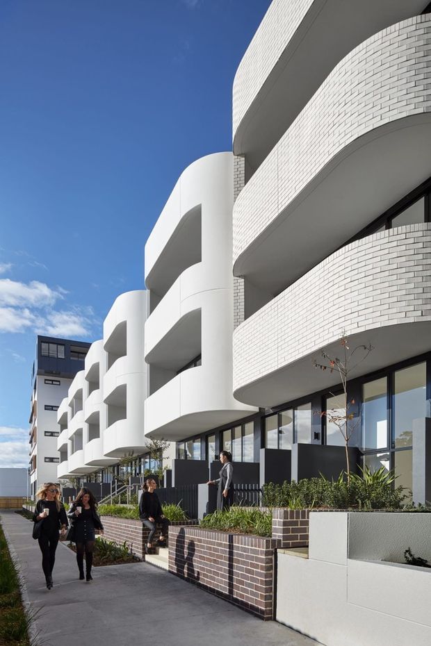 Exterior architectural photograph of a contemporary multi-storey apartment building viewed from a pedestrian walkway. The facade combines curved white rendered balcony forms with sweeping bands of light brickwork, dark-framed glazing, landscaped raised garden beds, and a strong linear repetition of balconies. Several people occupy the foreground walkway, with a clear blue sky behind the building.