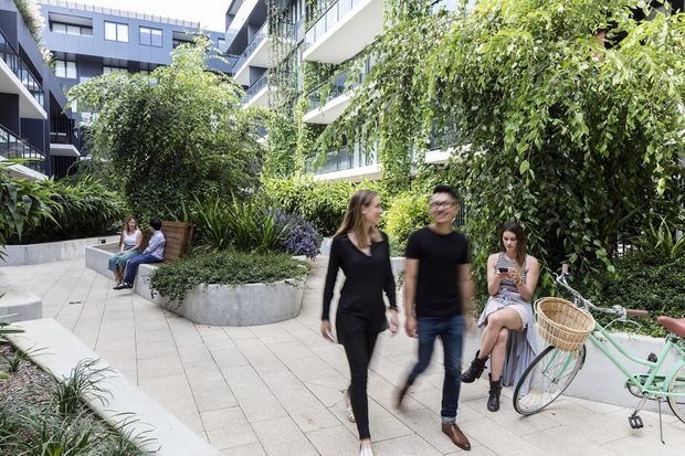 A landscaped communal courtyard and pedestrian walkway within a contemporary multi-storey residential development. Pale tiled paving winds between raised concrete planters filled with dense greenery, with integrated bench seating, residents, and a pastel bicycle. Modern dark and white apartment facades with balconies and extensive vertical planting enclose the space.