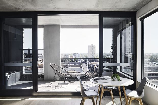 A bright contemporary apartment dining and balcony setting with a round white dining table and modern chairs beside full-height black-framed glazing. The open balcony has wire-frame lounge chairs and expansive urban views, while exposed concrete ceilings and columns create an industrial architectural character.