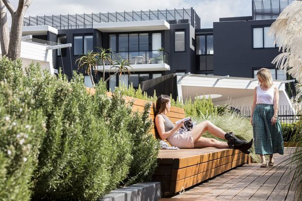 A contemporary multi-level residential building with a dark charcoal facade, extensive glazing, rooftop screening and balconies, viewed from a landscaped outdoor communal area. Dense planting surrounds timber-clad built-in bench seating and a paved walkway, with people relaxing in the foreground.