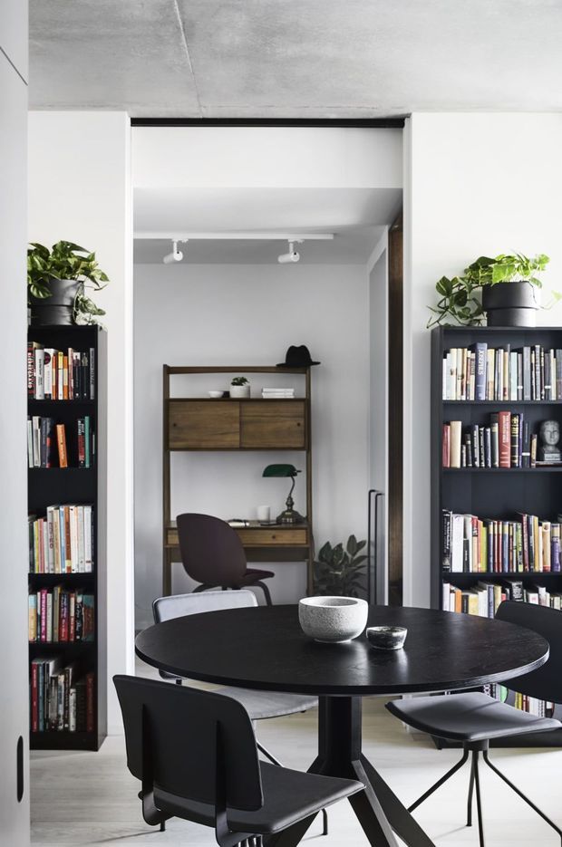 A refined open-plan dining and study area with a round black dining table and contemporary black chairs in the foreground. Full-height dark bookcases with plants and books flank a recessed home office, which contains a timber desk and shelving, task lamp, and decorative objects. The space has white walls, a pale concrete ceiling, light flooring, and dark timber and black accents.