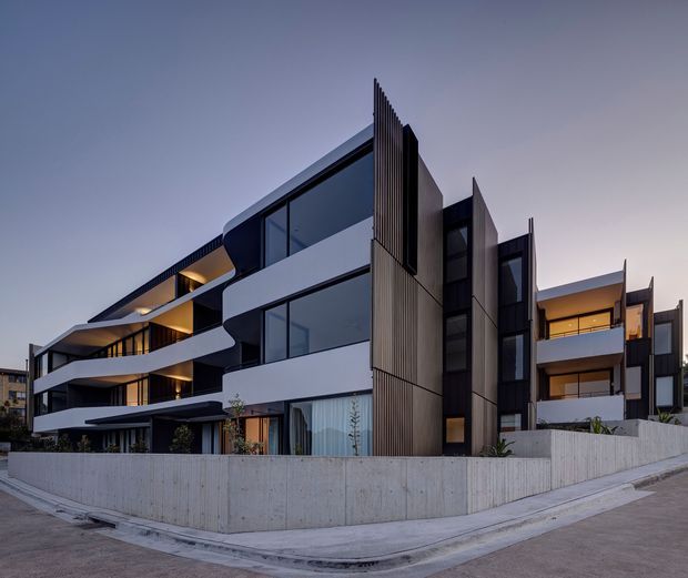 Exterior dusk view of a contemporary multi-storey residential apartment building with sculptural curved white balcony bands, extensive dark glazing, vertical ribbed facade screens, and exposed concrete boundary walls. Warm interior lighting contrasts with the cool blue-grey evening light.