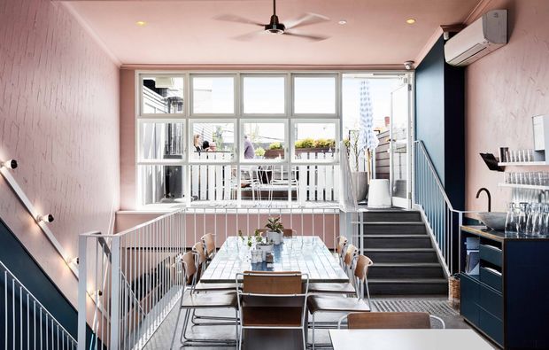 A stylish split-level dining and entertaining area with a long dining table, contemporary chairs, built-in dark cabinetry and bar storage, and stairs leading to an upper balcony or terrace. The space features pale pink textured walls, dark blue accents, extensive white-framed glazing, a ceiling fan, and bright natural light. A small outdoor balcony with seating and plants is visible beyond the windows.