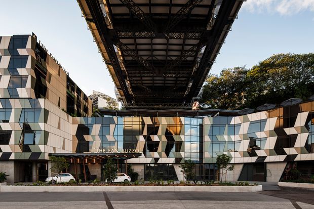 Exterior architectural view of a contemporary multi-storey commercial building beneath a large elevated steel structure or railway viaduct. The facade combines geometric white, grey, and dark panels with extensive reflective glazing, creating a faceted pattern. A landscaped forecourt and driveway sit in front, with mature trees and blue sky in the background.