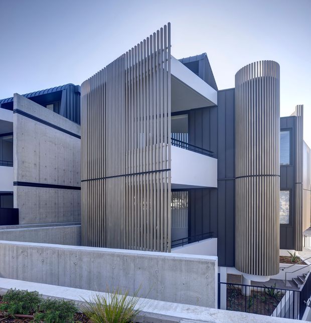 Contemporary multi-storey residential facade featuring prominent curved vertical slatted screens, dark metal cladding, white projecting balconies and broad exposed concrete walls. The building is photographed from an elevated exterior viewpoint, with layered terraces, railings and landscaped planting visible in the foreground.