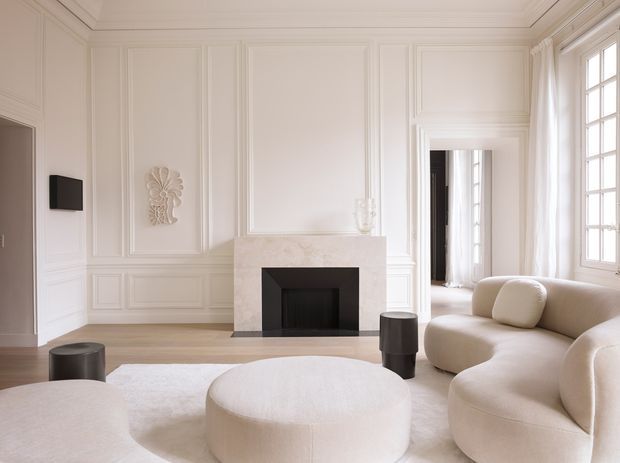 Bright, refined living room with extensive white decorative wall moulding and panelled detailing, a pale stone fireplace with black inset, tall multi-pane windows, and a sculptural cream upholstered sectional arranged around a large round ottoman. The room has a restrained monochromatic palette and elegant contemporary-classic styling.