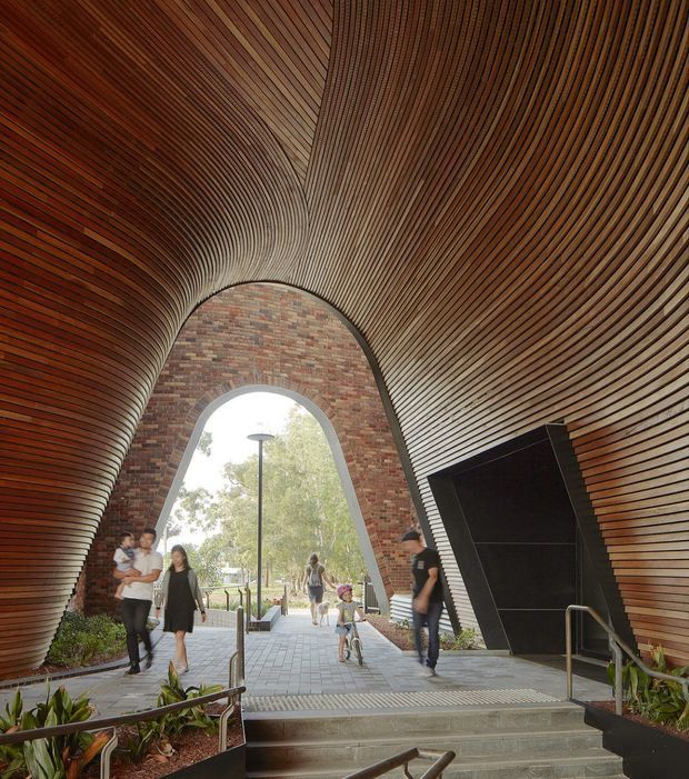 A dramatic vaulted pedestrian passage with a high arched opening, lined throughout with closely spaced curved timber slats in warm reddish-brown tones. Exposed brickwork frames the opening, while concrete steps, metal handrails, planted beds, and people animate the public circulation space. Daylight and trees are visible beyond the arch.