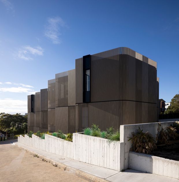 Exterior view of a contemporary multi-level building on a sloped suburban site. The facade is dominated by closely spaced vertical dark metal fins, with stepped massing, narrow vertical glazing, and curved roofline detailing. Pale concrete retaining walls and landscaped planters line the driveway and sidewalk.