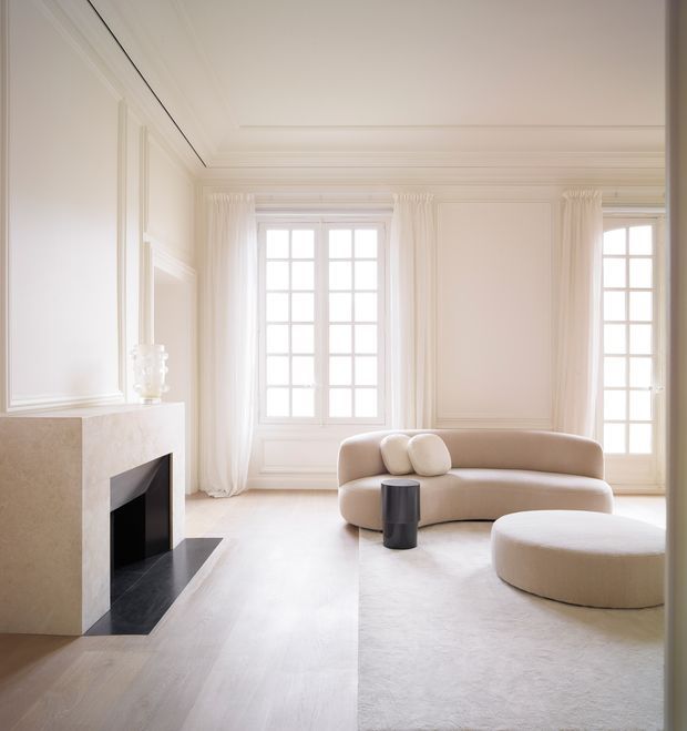 A bright, refined living room with white paneled walls, ornate ceiling detailing, tall multi-pane windows with sheer curtains, a minimalist stone fireplace, and a curved cream sofa with a matching round ottoman. Pale timber flooring, a light rug, and restrained furnishings create an airy, sophisticated interior.