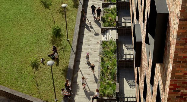 Aerial oblique view of a landscaped urban pedestrian courtyard or walkway beside a multi-storey brick building. The paved path is lined with young trees, planted garden beds, slim contemporary light poles, balconies, and projecting dark-framed windows or balconies. Several pedestrians are walking through the space, with a broad lawn visible on the left.
