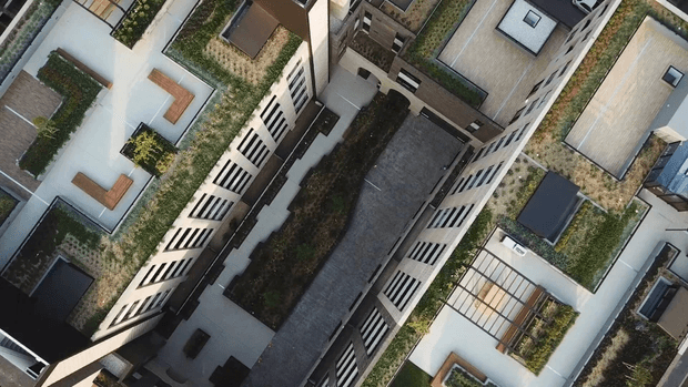 Aerial view of a dense contemporary multi-storey building complex with flat roofs, rooftop gardens, landscaped terraces, paved courtyards, and patterned facade screens. The image focuses on the building forms and green roof infrastructure rather than a single interior space.
