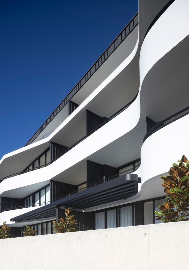 Upward-looking architectural exterior photograph of a multi-storey contemporary building with sweeping white curved balcony slabs, dark metal balustrades, black-framed glazing, and projecting dark horizontal awnings. A pale concrete boundary wall and small planted trees occupy the foreground against a clear deep-blue sky.